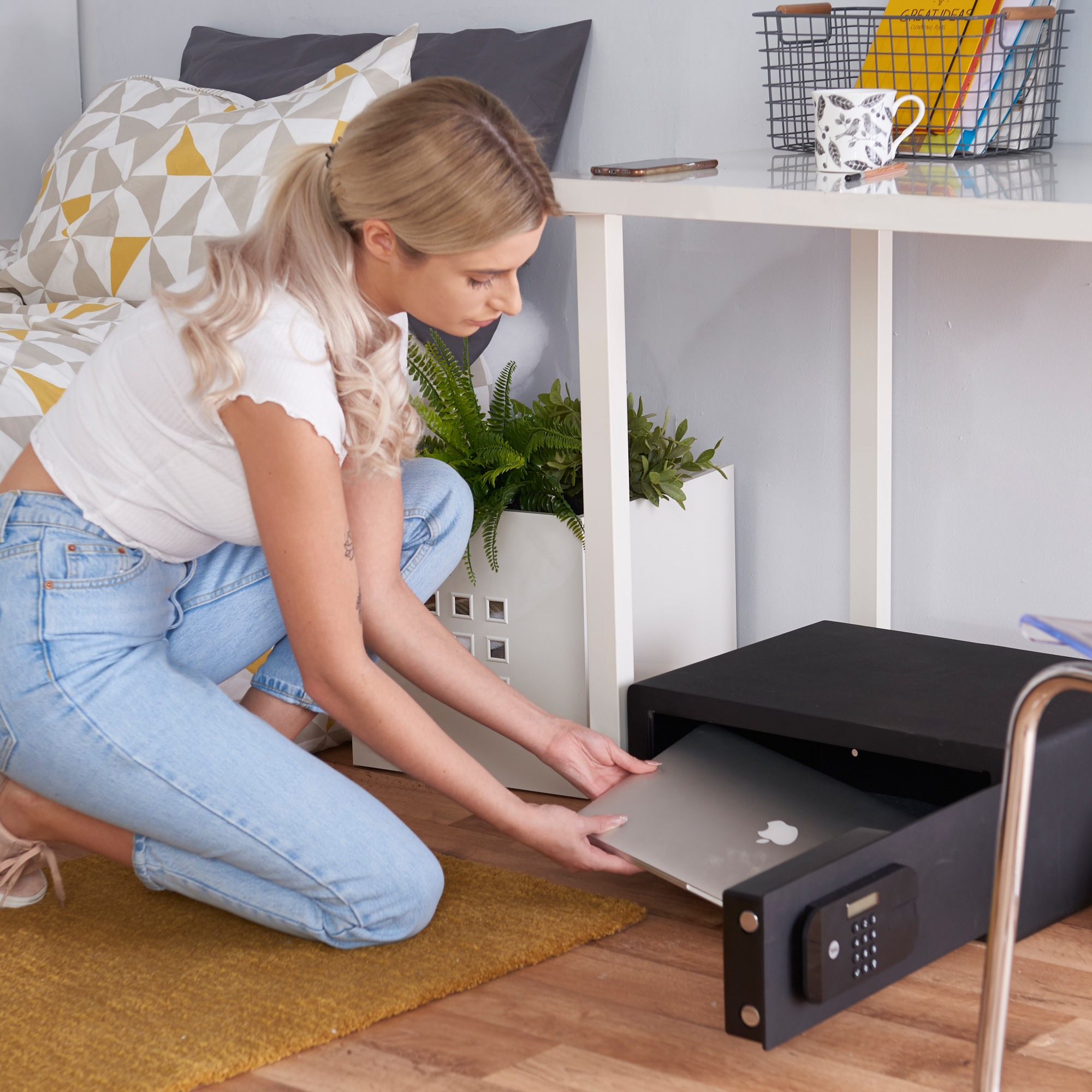 Eine Frau in Jeans und weißem Oberteil legt einen Laptop in einen schwarzen Safe unter einem Schreibtisch.