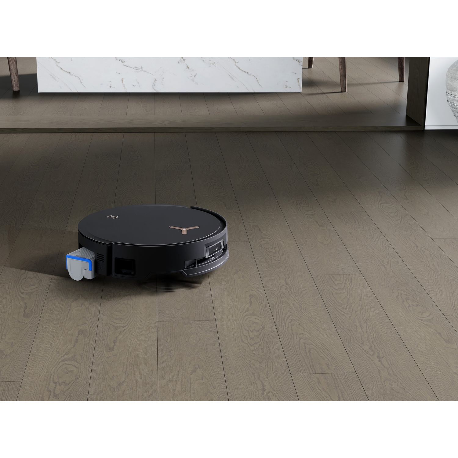 Un aspirapolvere robotico nero su un pavimento di legno. Lo sfondo include un piano cucina e un vaso.