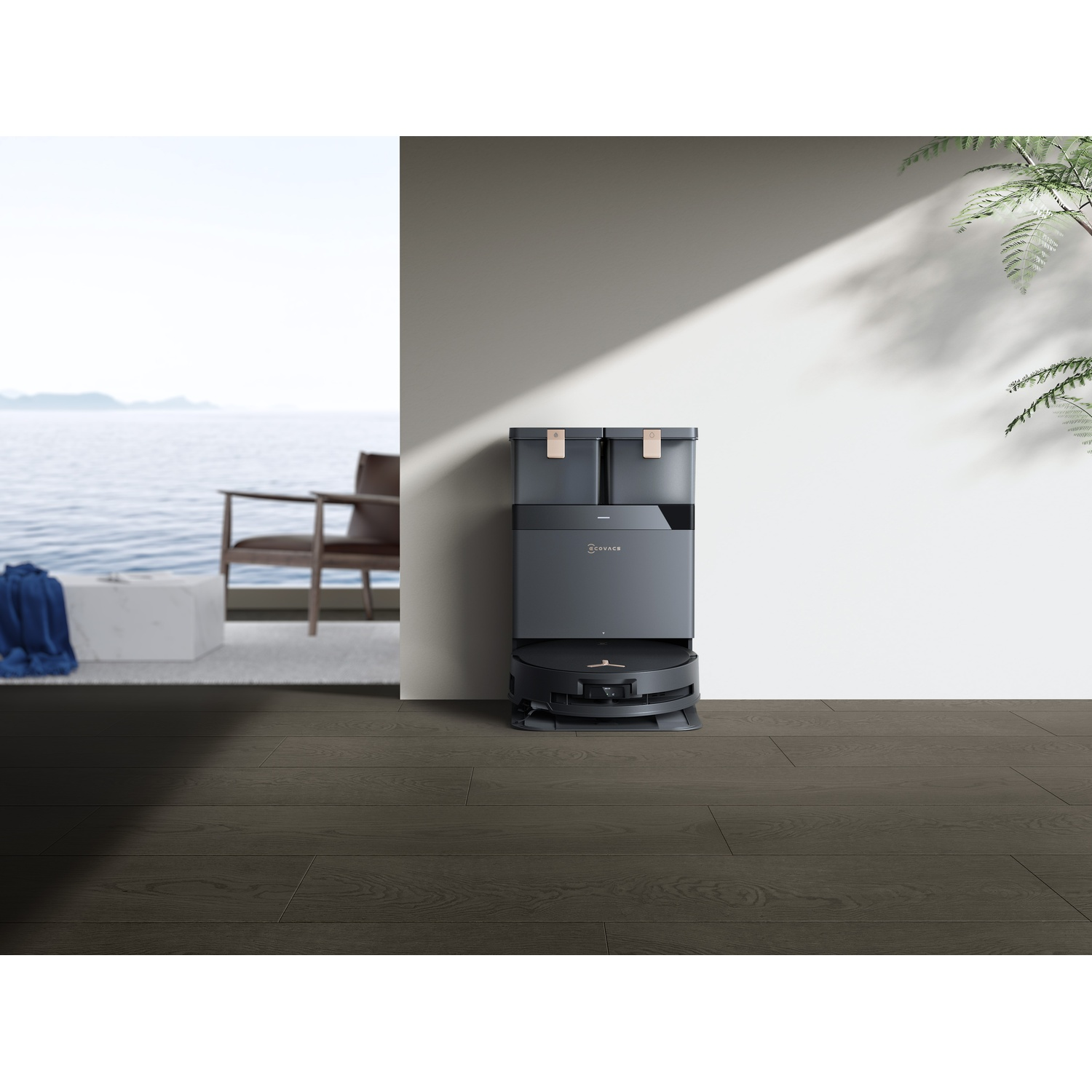 Un aspirapolvere robotico si trova su un pavimento in legno. Una stazione di pulizia è dietro di esso. Una sedia e l'oceano sono sullo sfondo.