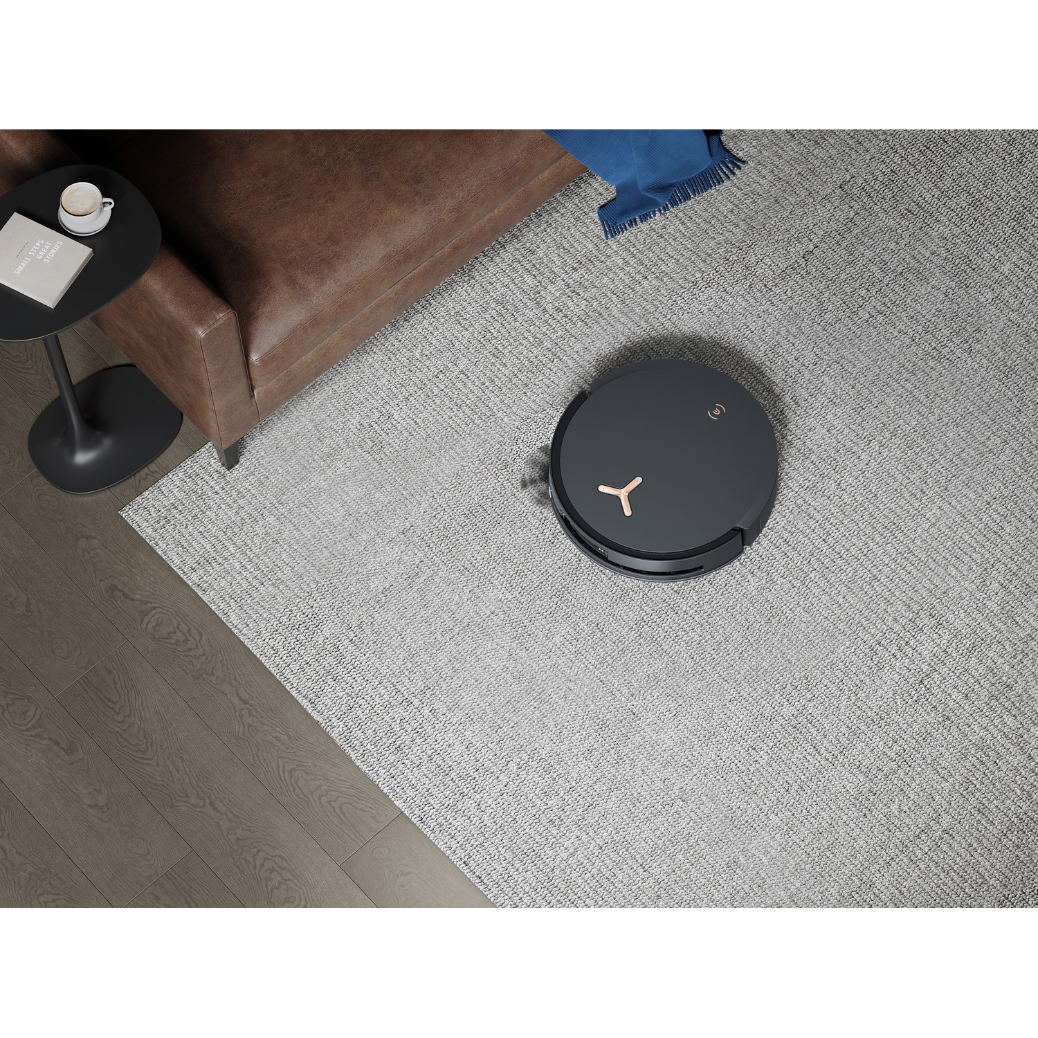 Un aspirapolvere robotico su un tappeto grigio. Sono visibili anche un divano marrone, un tavolino con una tazza di caffè e una coperta blu.