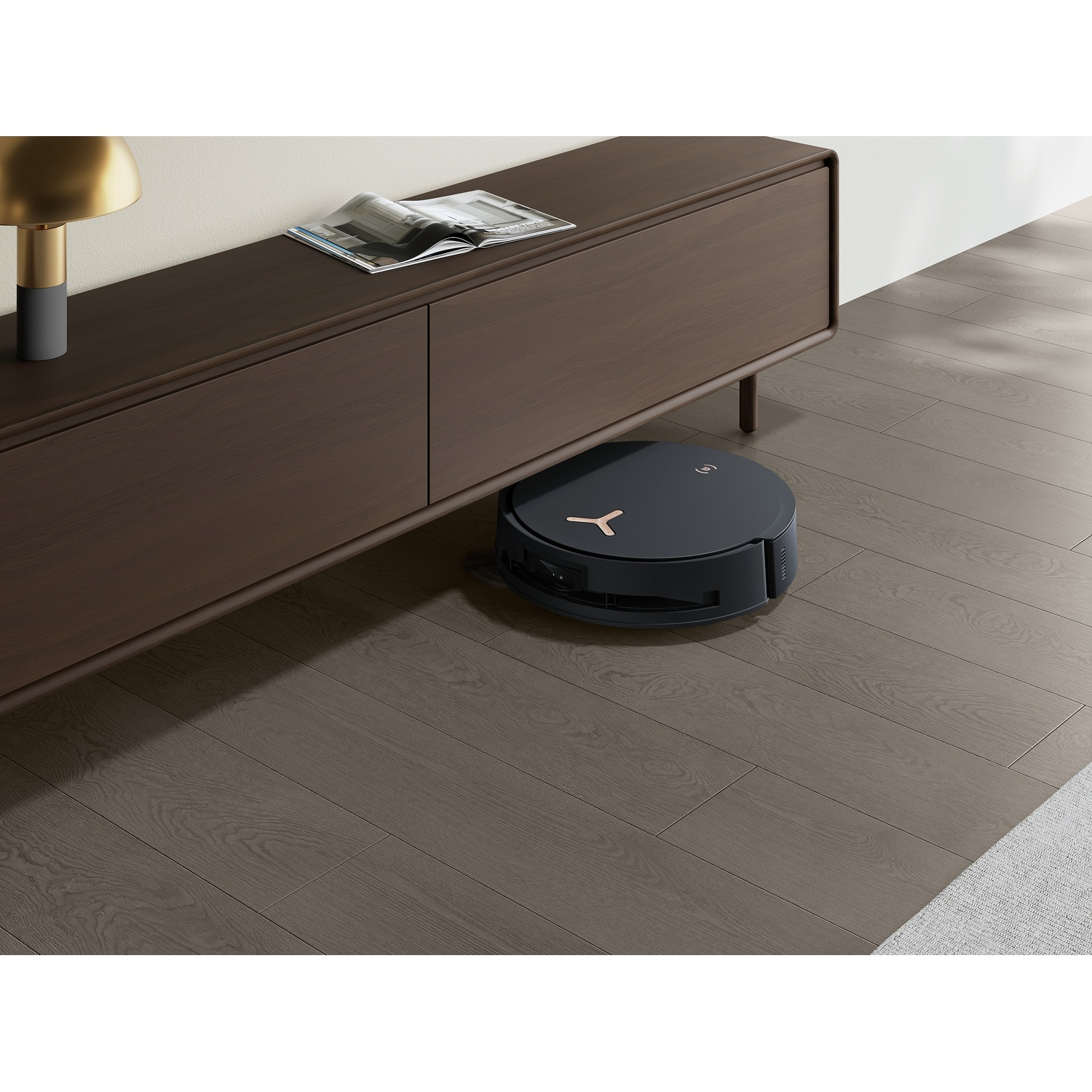 Un aspirapolvere robotico nero è su un pavimento in legno. Un mobile marrone scuro con una rivista e una lampada è sullo sfondo.