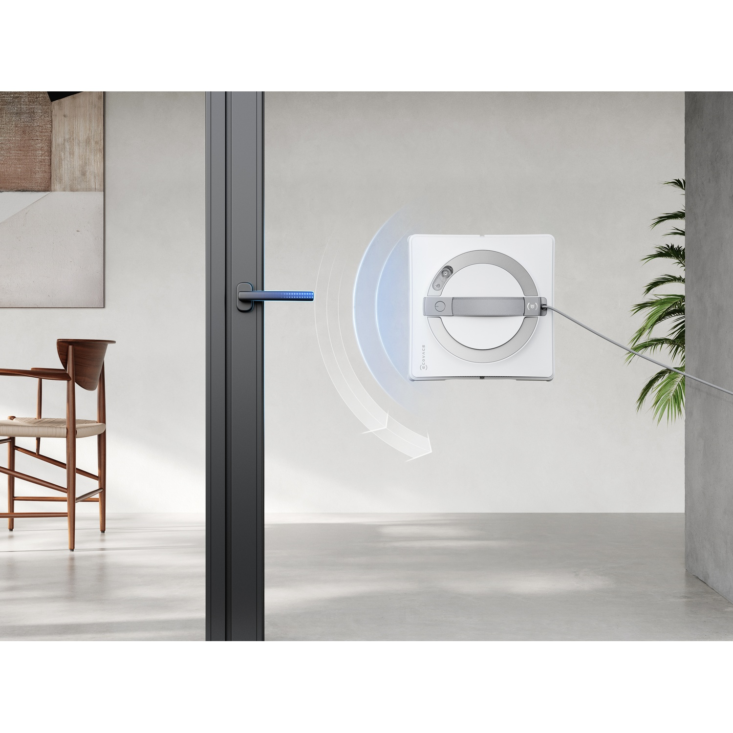 Un robot pulisci vetri bianco pulisce una porta a vetri. A sinistra e a destra ci sono una sedia di legno e una pianta. È visibile una maniglia.
