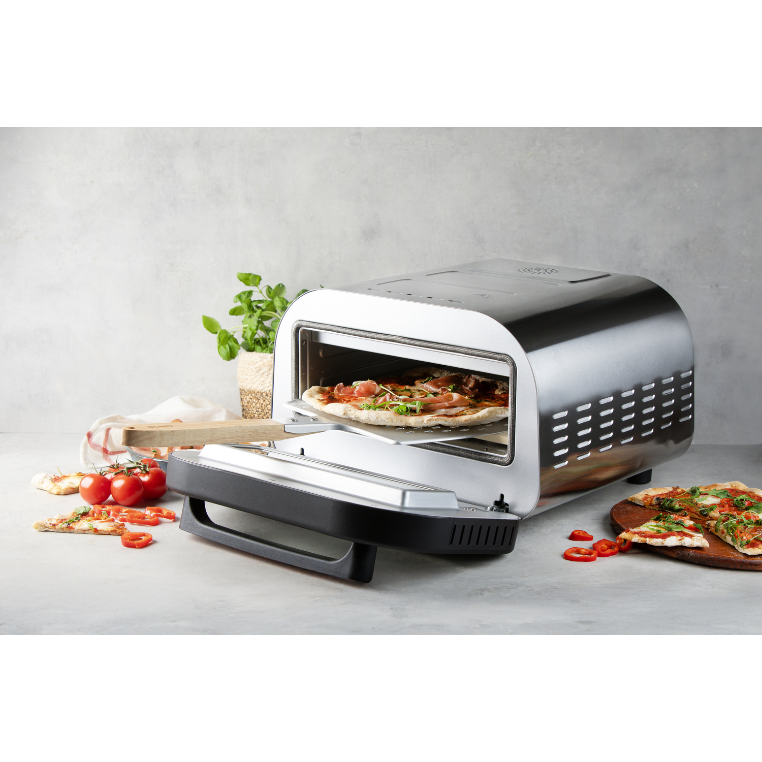 A silver toaster oven with a pizza inside, pizza slices, tomatoes, and a plant sit on a table.