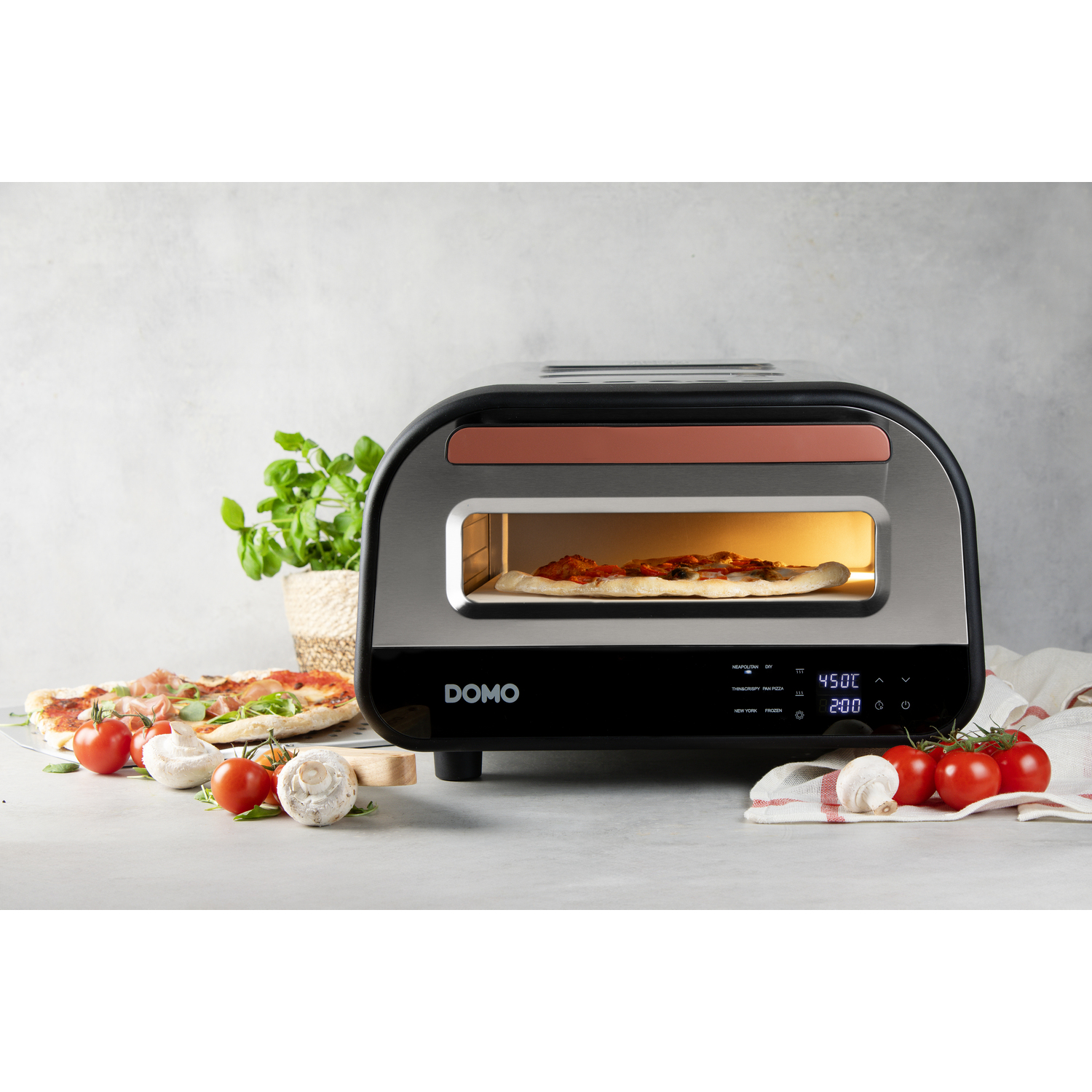 A pizza oven with a pizza inside, tomatoes, mushrooms, and a pizza on the left. A plant is in the background.