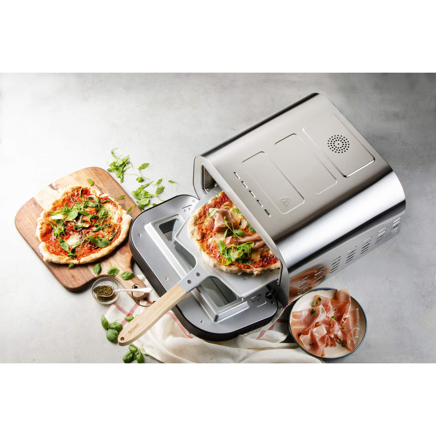 A pizza oven with two pizzas, one on a wooden board, the other in the oven. Slices of ham are on a plate.