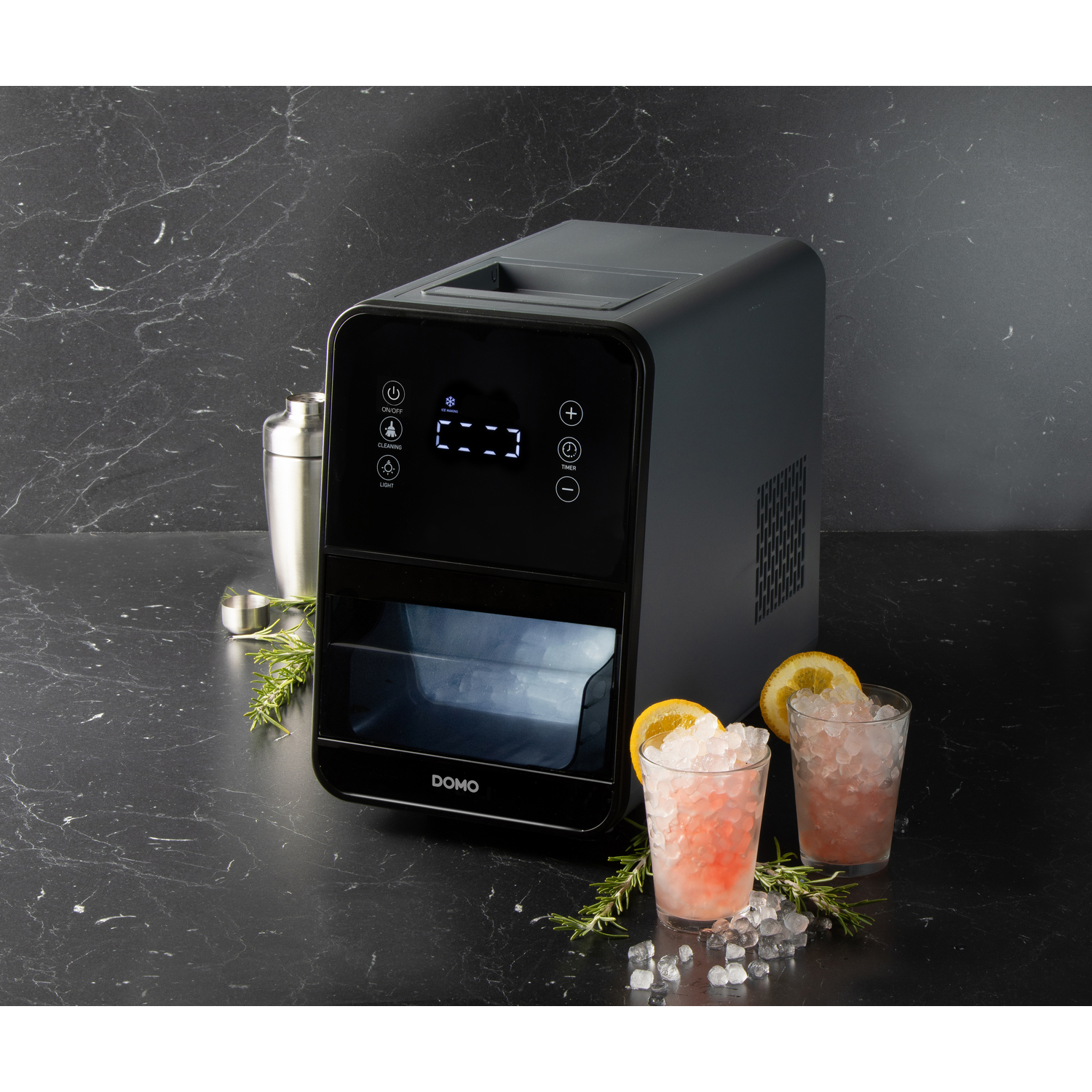 An ice maker, drinks, and bar tools on a black surface. Ice maker is black. Drinks have orange slices. Cocktail shaker.