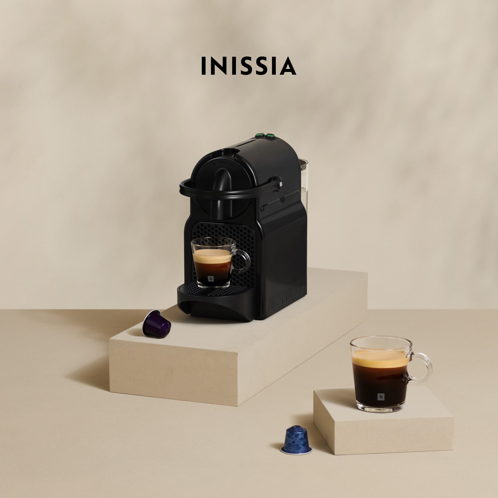 Czarny ekspres Nespresso Inissia z kawą w szklance. Dwie kapsułki i filiżanka kawy na beżowych blokach.