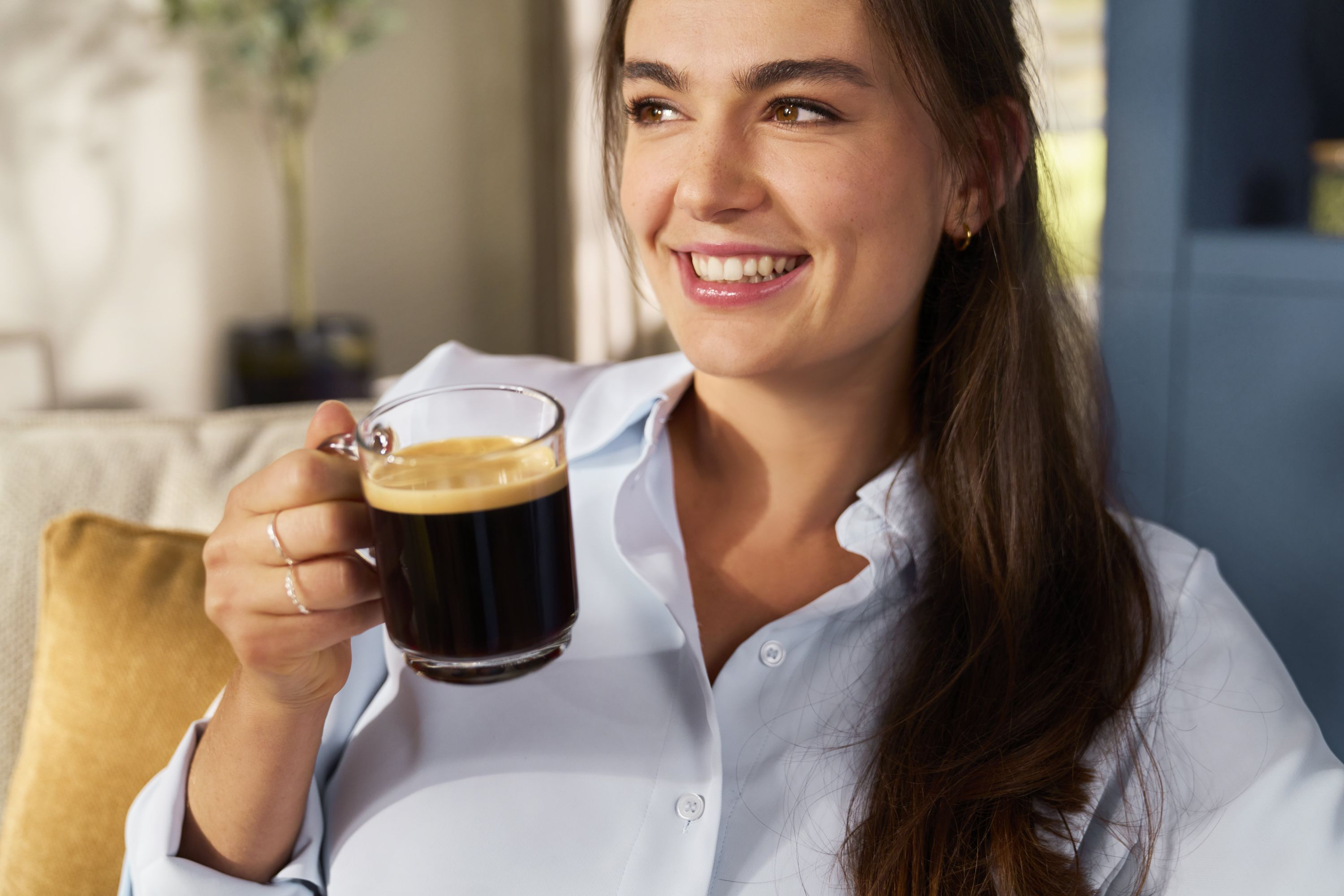Mujer sonríe sosteniendo café. Lleva camisa celeste. Fondo: sofá y planta.