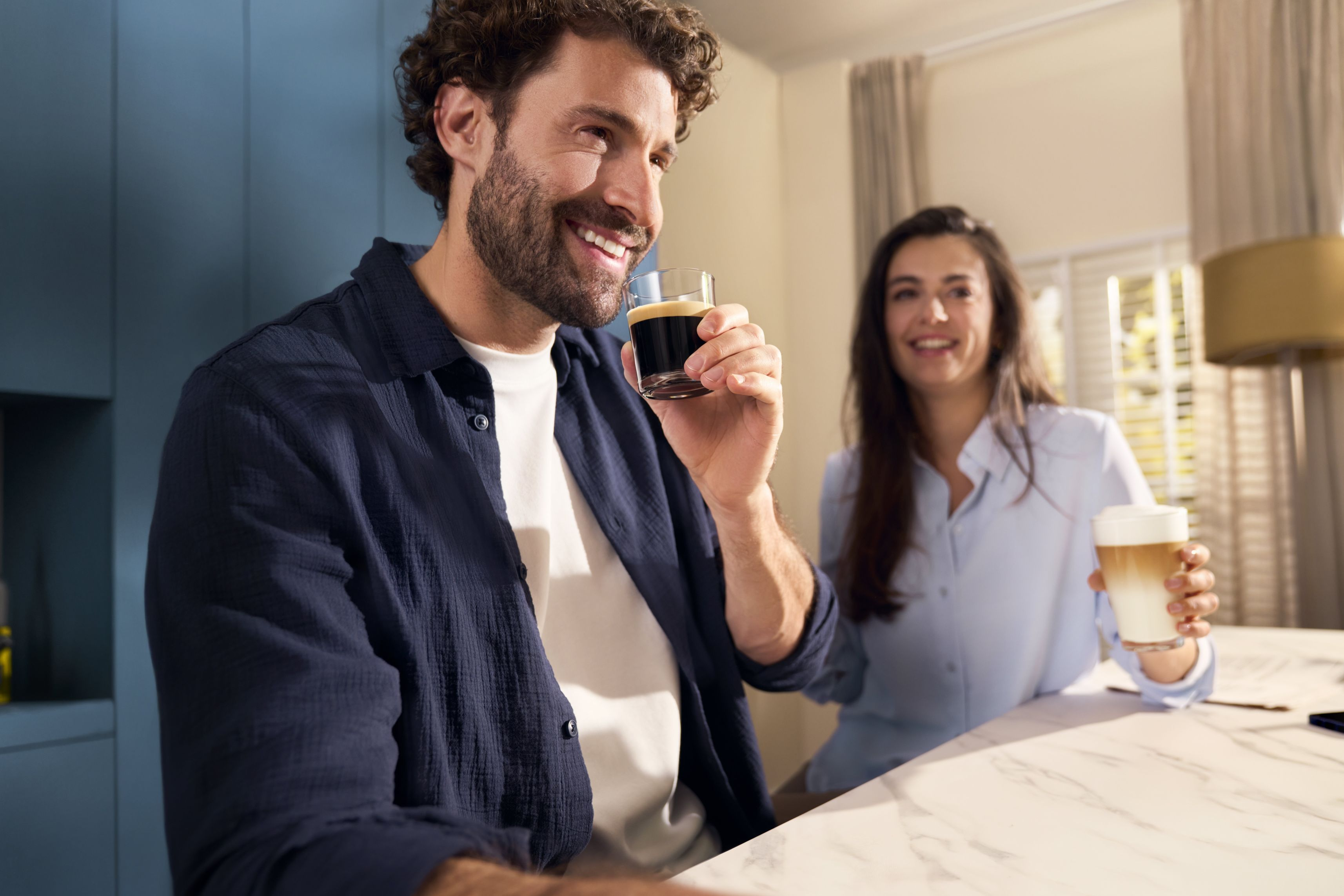 Un hombre bebe café, sonriendo. Una mujer sostiene un latte. La escena tiene un ambiente ligero y aireado.