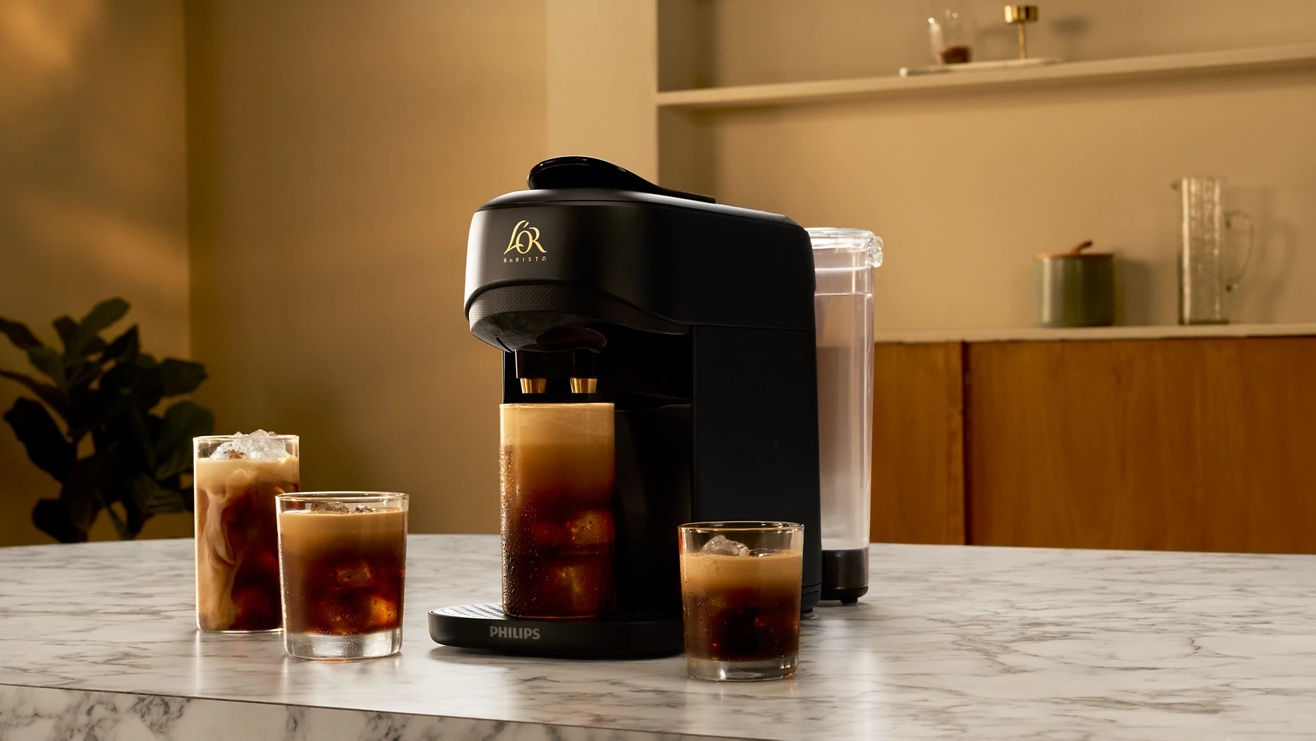 Una cafetera negra con tres vasos de café helado sobre una encimera de mármol. El fondo muestra una cocina.