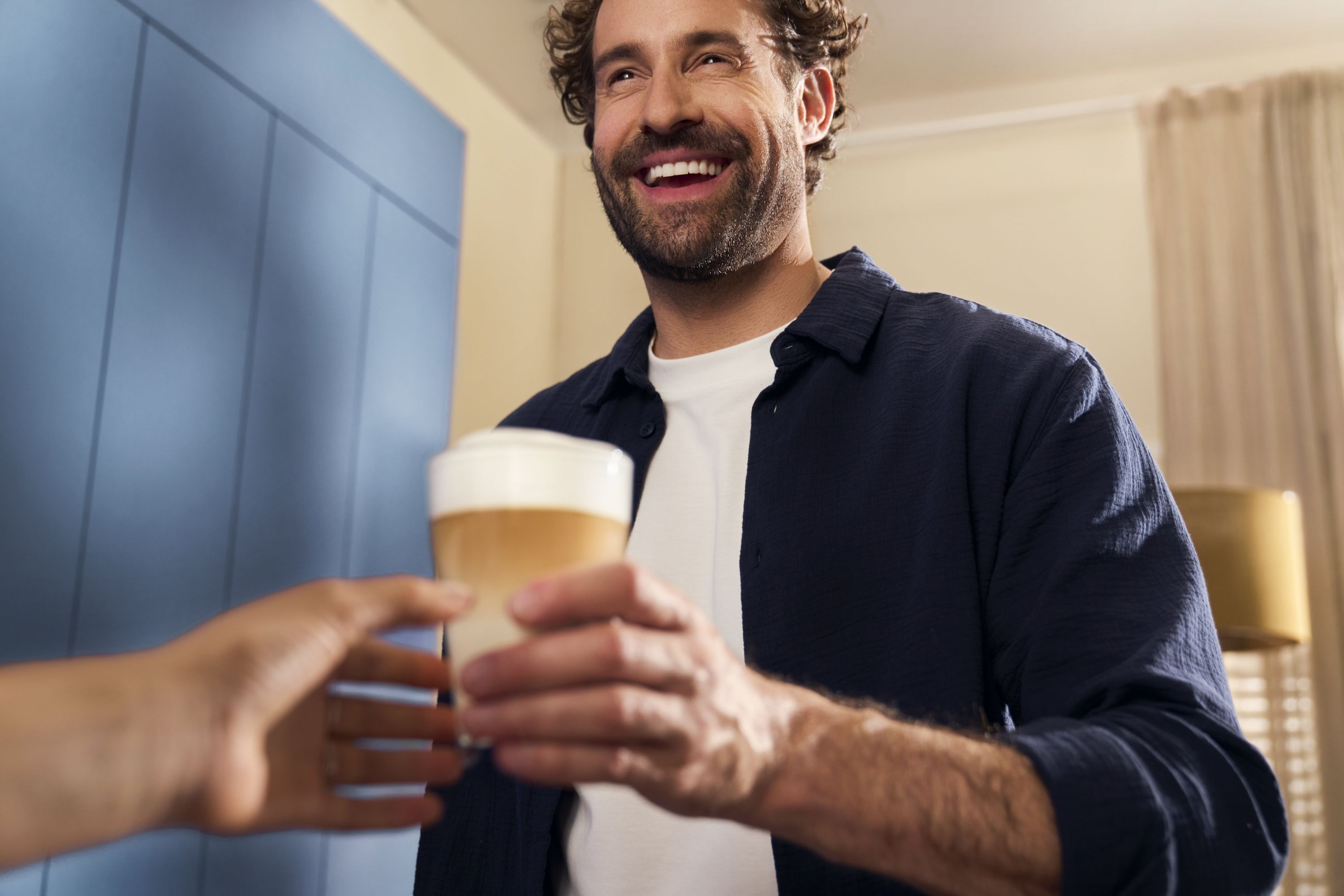 Un hombre sonríe ofreciendo un latte. Una mano alcanza la bebida. Pared azul al fondo.