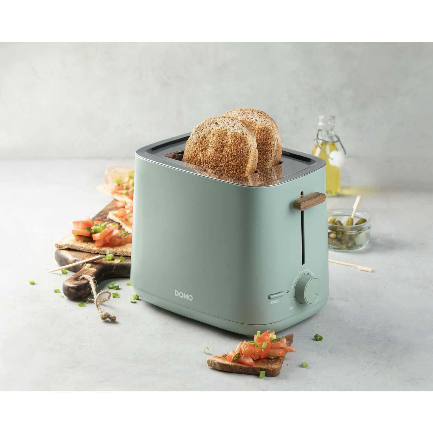 A teal toaster with two slices of toast. Beside it are bread slices with toppings, a jar of pickles, and oil.