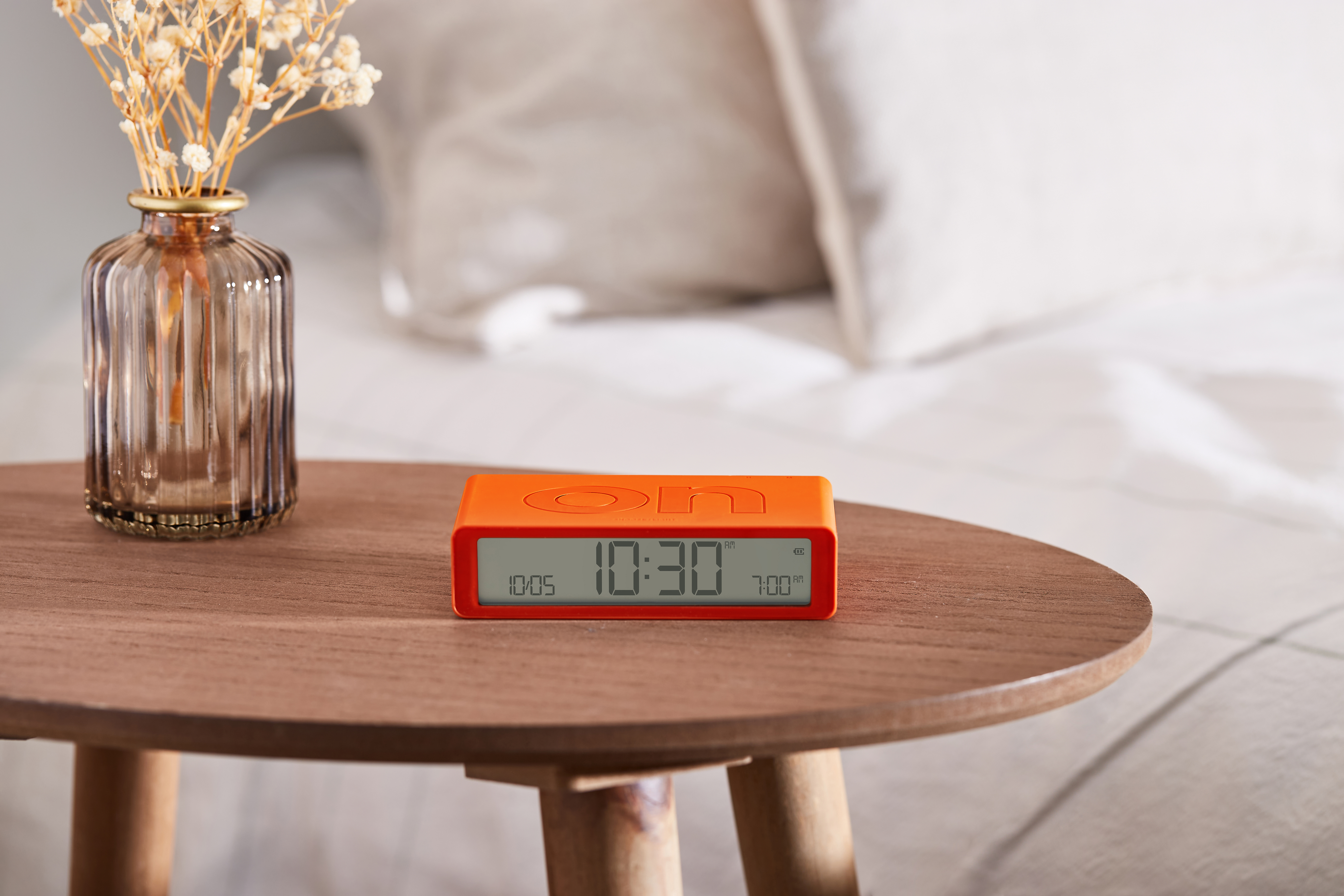 Ein orangefarbener Wecker zeigt 10:30 Uhr auf einem Holztisch an. Daneben steht eine Vase mit Trockenblumen, im Hintergrund ein Bett.