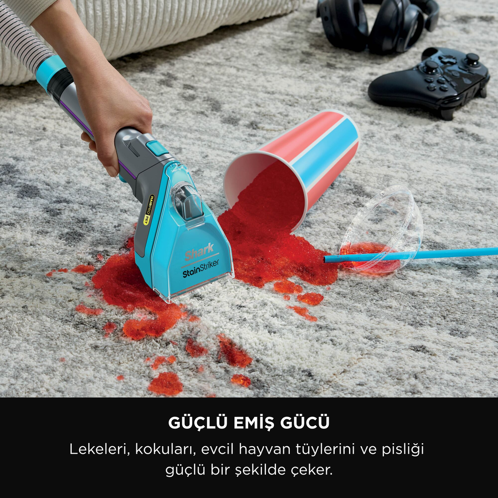 Bir Shark StainStriker halıdaki kırmızı bir dökülmeyi temizliyor. Dökülen içecek, kulaklıklar ve bir oyun kumandası arka planda.