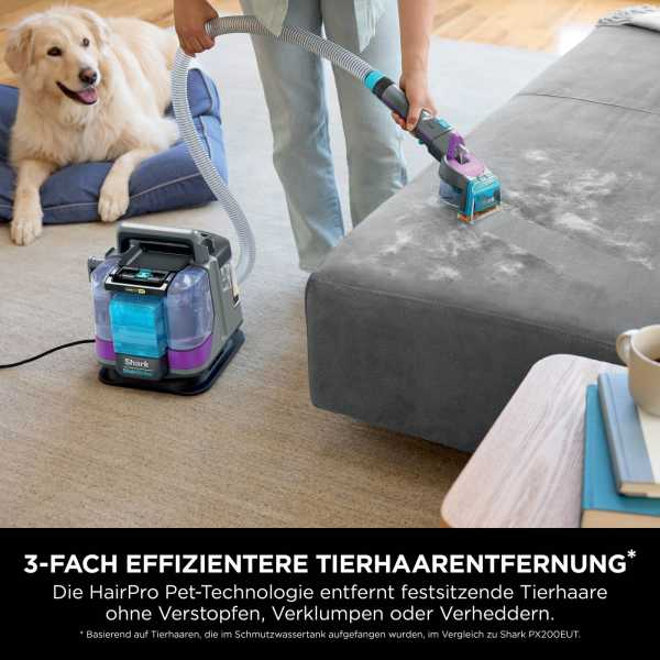 Eine Person saugt ein graues Sofa mit einem Shark-Reiniger. Ein Hund ruht auf einem blauen Kissen. Ein Tisch hält Bücher und eine Tasse.