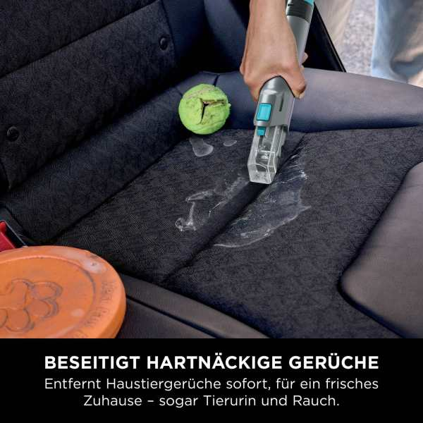 Eine Hand besprüht einen Autositz mit einem Reinigungswerkzeug. Ein grüner Ball und eine orangefarbene Scheibe befinden sich in der Nähe. Reinigungsaktion auf dunklem Sitz.