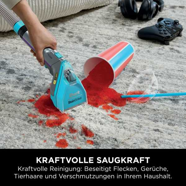 Ein Teppich ist mit rotem Pulver befleckt. Eine Person benutzt einen blau-grauen Staubsauger, um ihn zu reinigen. Eine Tasse ist umgestürzt.