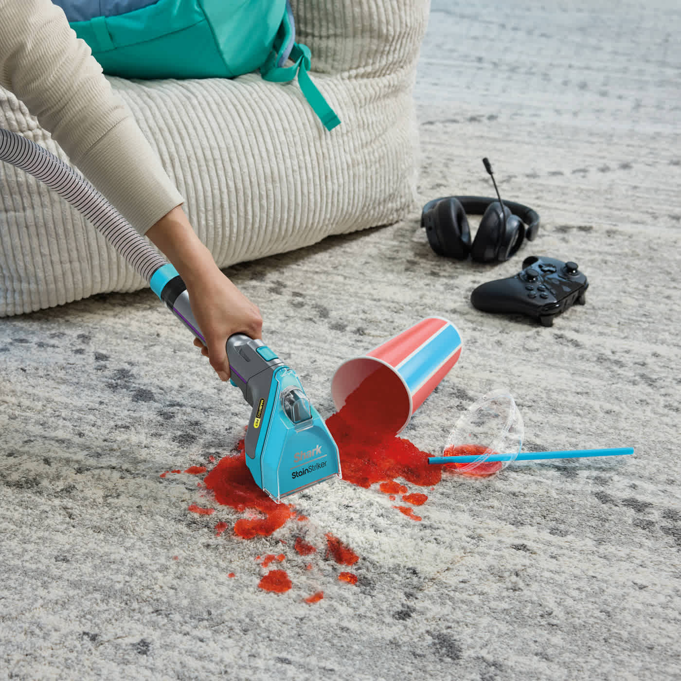 Ein verschüttetes rotes Getränk wird mit einem blauen Shark StainStriker gereinigt. Becher, Strohhalm, Gaming-Headset und Controller sind vorhanden.