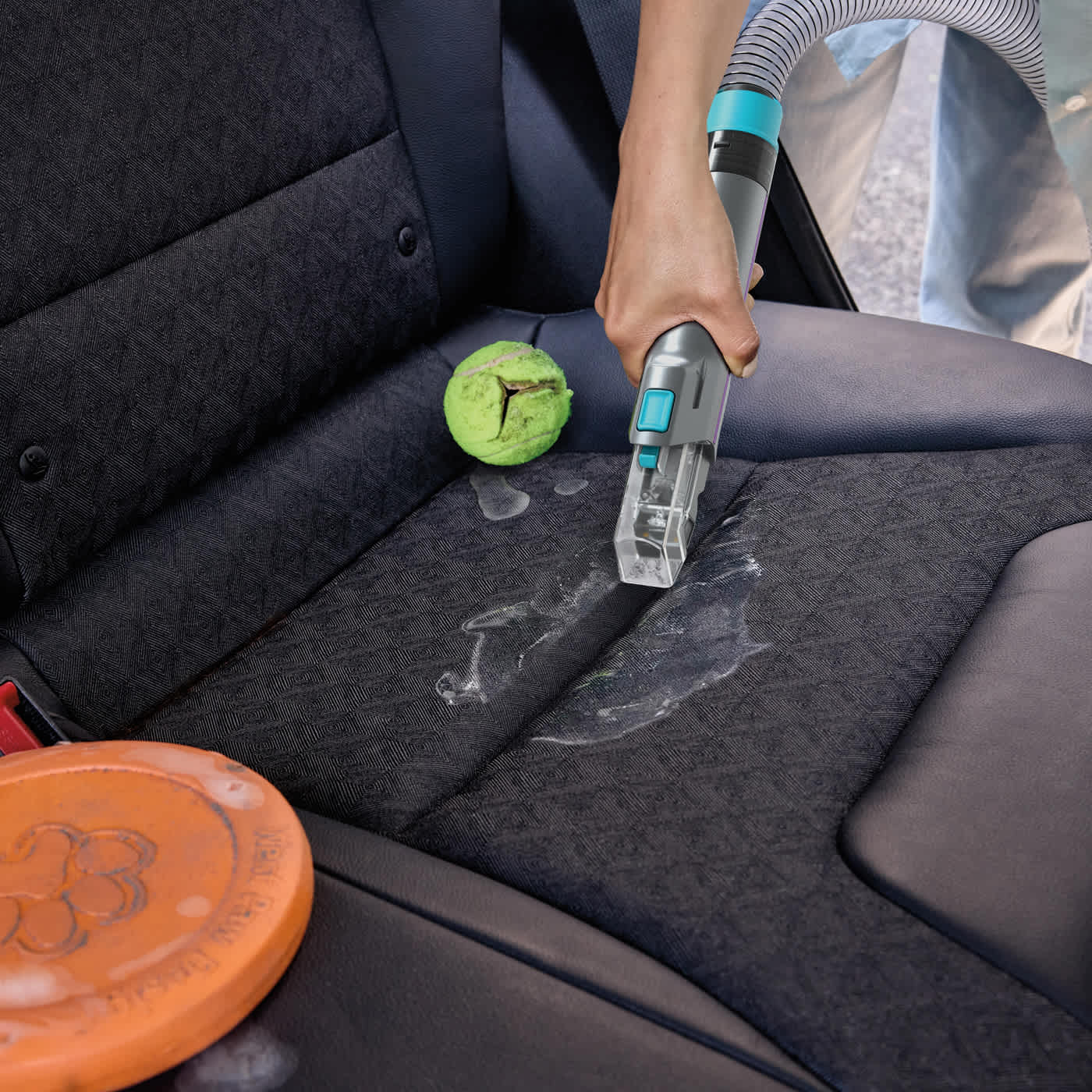 Eine Person reinigt einen Autositz mit einem Staubsauger. Ein Tennisball und eine orangefarbene Frisbeescheibe liegen auf dem Sitz. Der Sitz ist schwarz.