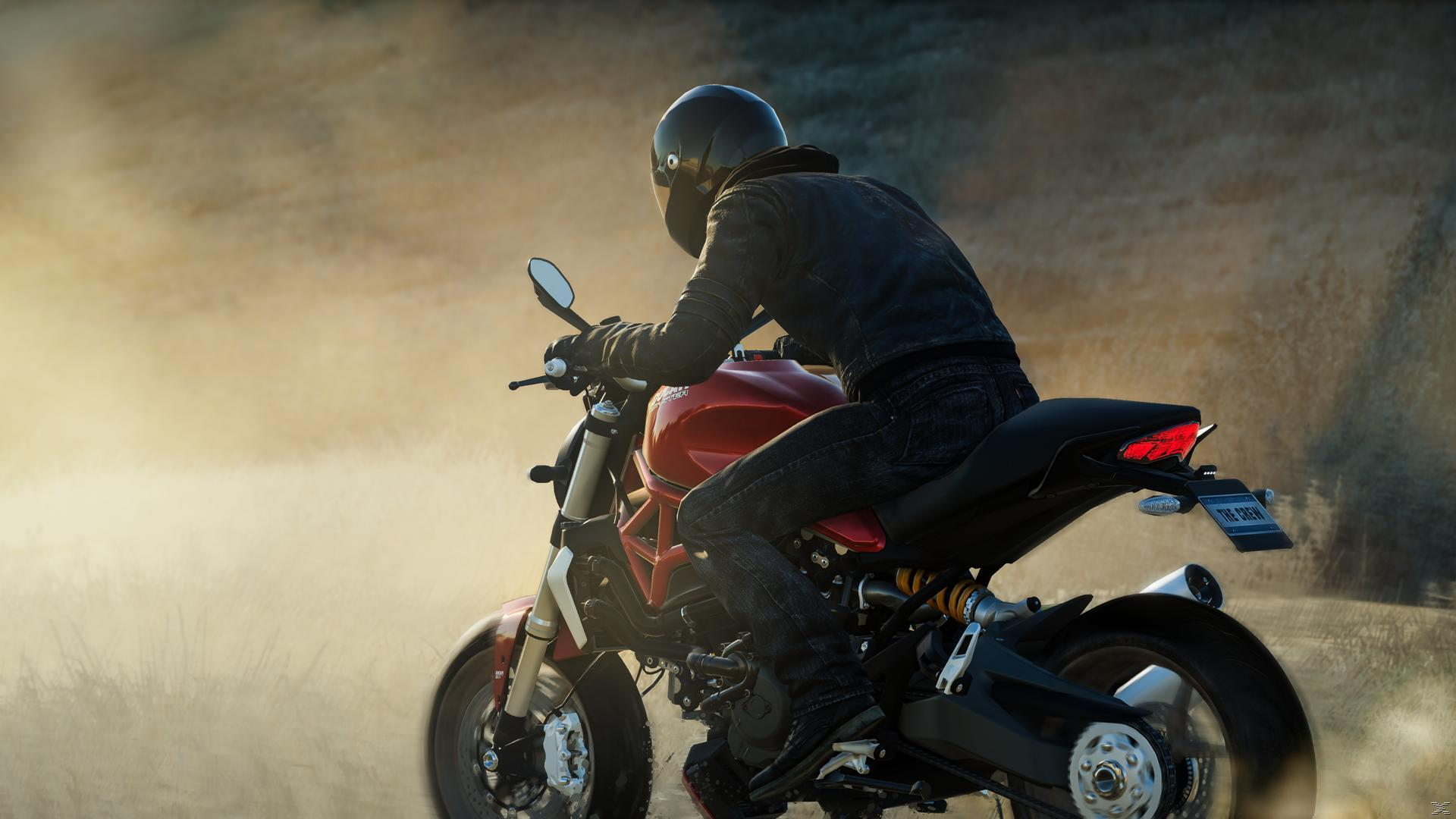 Eine Person auf einem roten Motorrad, mit schwarzem Helm und Jacke, fährt auf einer staubigen Straße. Der Hintergrund ist unscharf.
