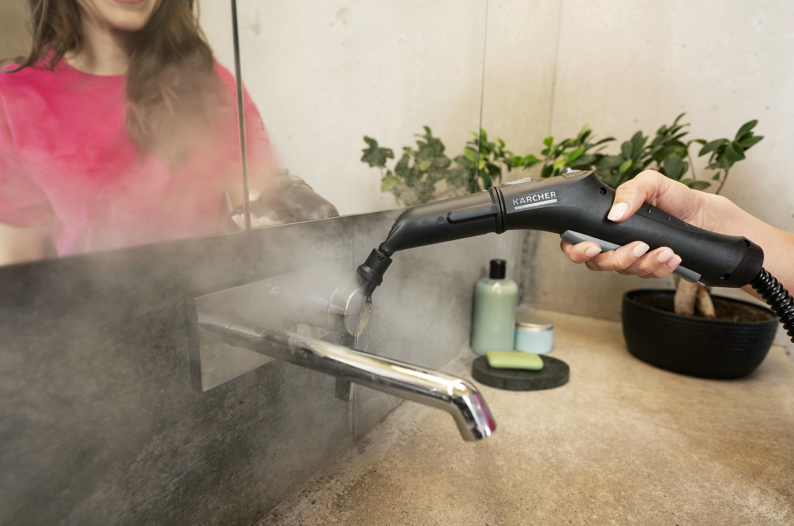 Egy személy Karcher gőztisztítót használ a fürdőszoba csapjához. Gőz emelkedik. Egy nő tükörképe látható.