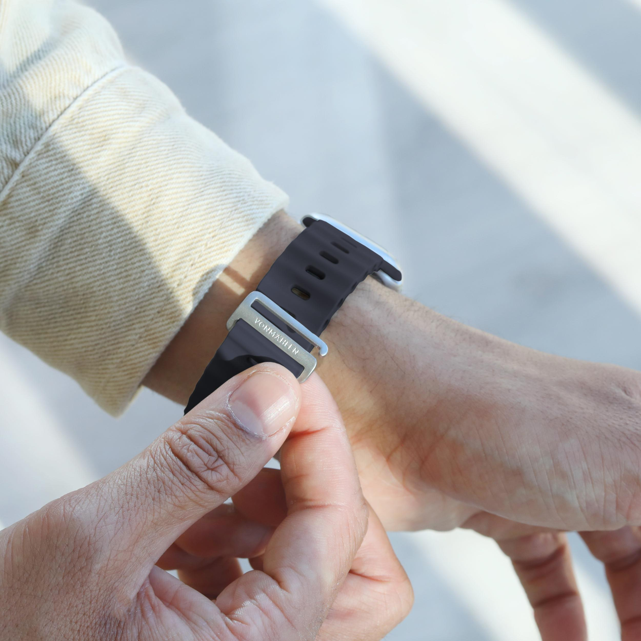 Eine Person befestigt eine schwarze Armbanduhr mit silberner Schließe. Die Uhr befindet sich am linken Handgelenk, ein beiger Jackenärmel ist sichtbar.