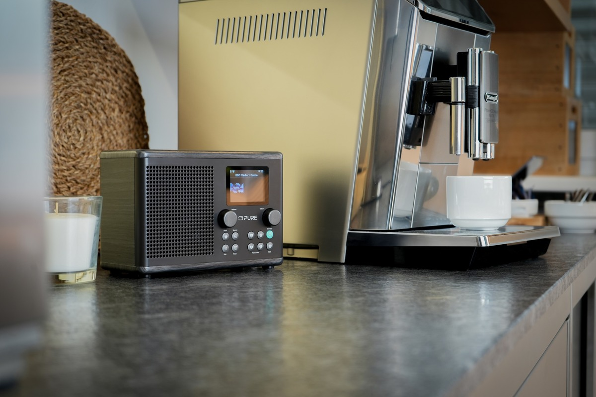 Une radio noire, une machine à café et une tasse blanche sur un comptoir. Une bougie et une décoration en osier sont également dans la scène.