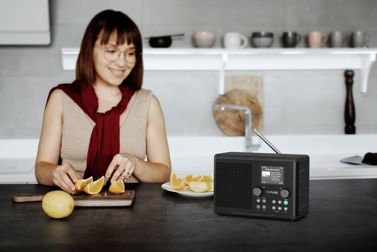 Une femme avec des lunettes coupe une orange. Une radio noire est sur un comptoir. Des oranges coupées et un citron sont à proximité.