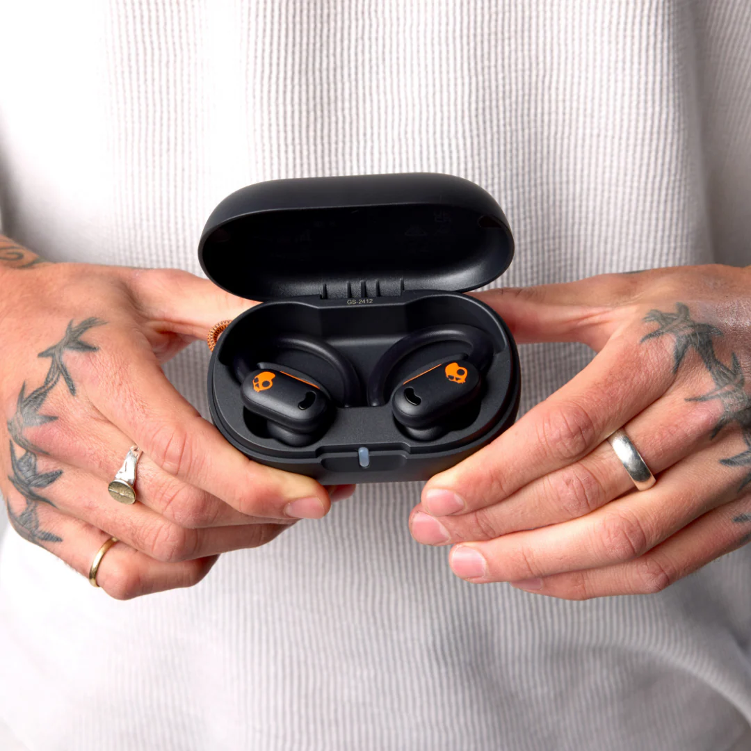 Person hält eine offene schwarze Kopfhörerhülle mit orangefarbenen Skullcandy-Ohrhörern. Hände haben Tätowierungen und Ringe. Weißes Hemd im Hintergrund.