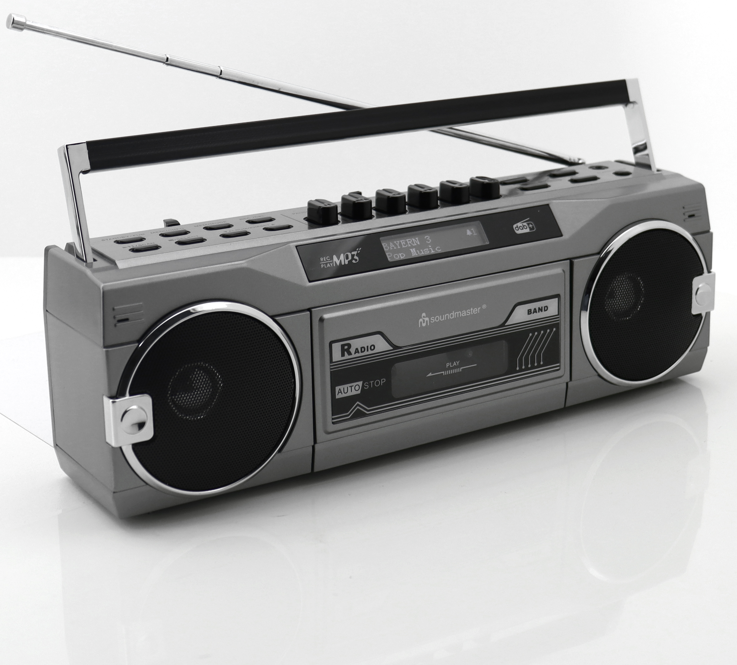 Grauer Ghettoblaster auf weißer Oberfläche. Er hat Lautsprecher, Knöpfe und einen Griff. Das Radio zeigt Senderinformationen.