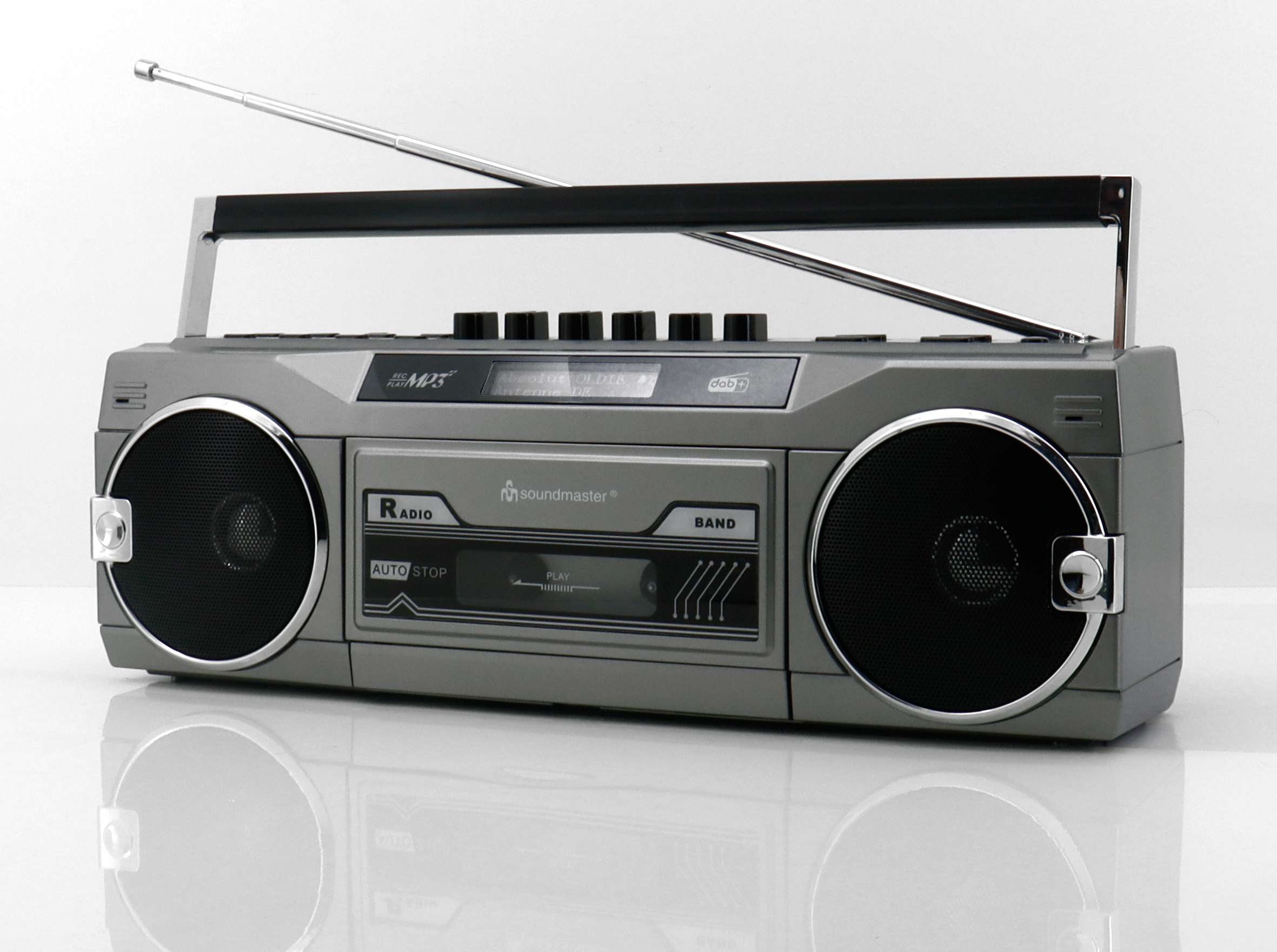 Grauer Ghettoblaster mit Lautsprechern, Antenne und Griff auf reflektierender Oberfläche vor weißem Hintergrund.