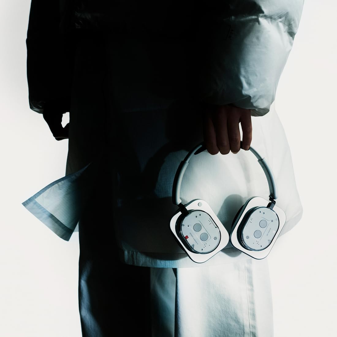 Une personne en vêtements blancs tient un casque blanc avec des oreillettes carrées. Fond sombre. Le bras de la personne est visible.