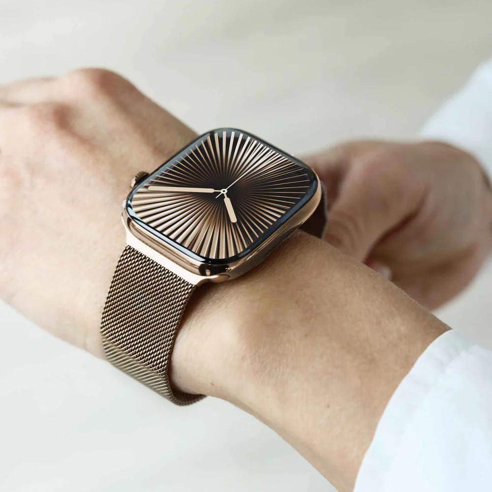 Handgelenk mit goldener Uhr. Die Uhr hat ein schwarzes und goldenes Zifferblatt und ein Mesh-Armband. Die Person trägt ein weißes Hemd.