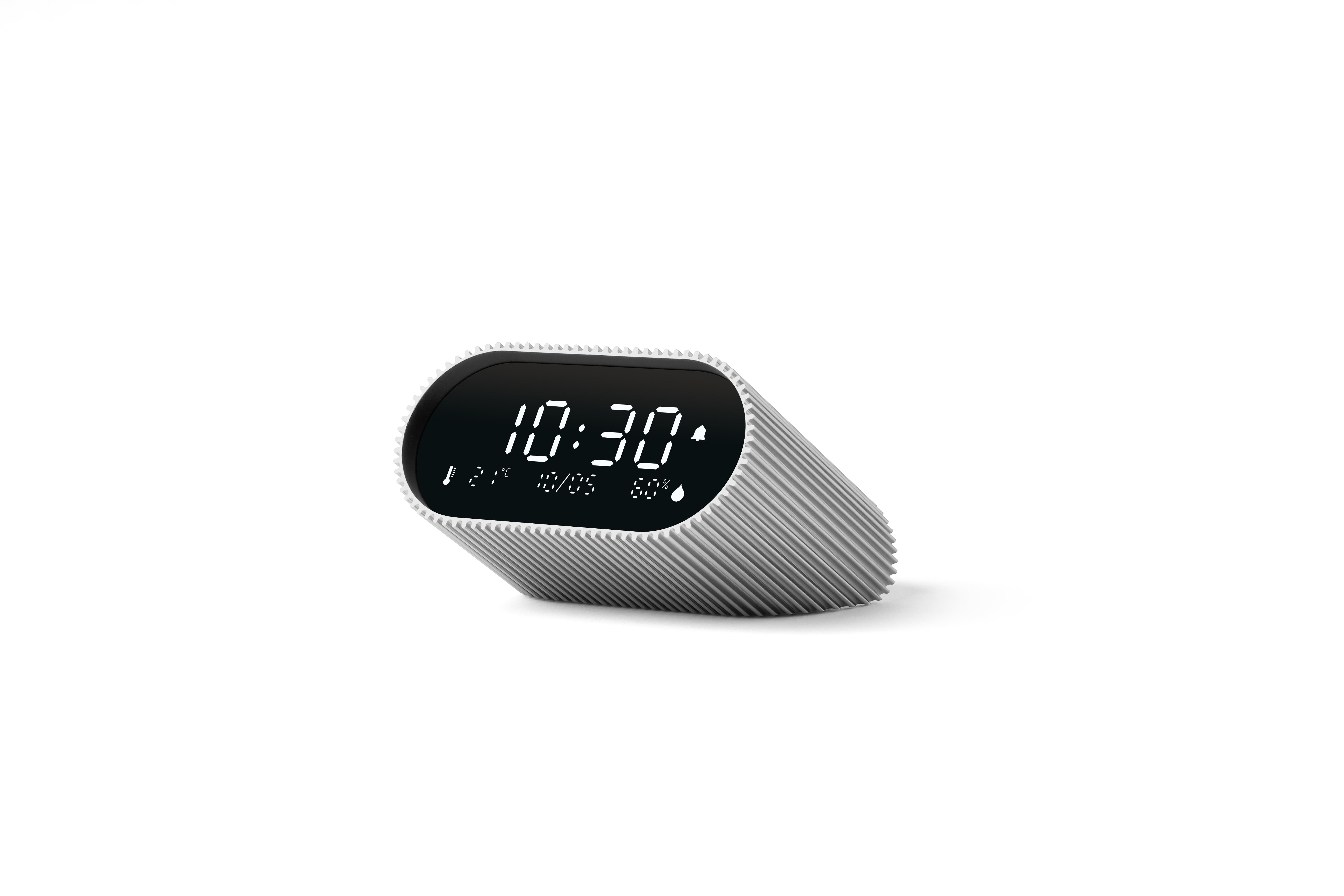 Ein digitaler Wecker zeigt 10:30 Uhr an. Er ist weiß mit schwarzem Bildschirm und gerippten Seiten, auf weißer Oberfläche.