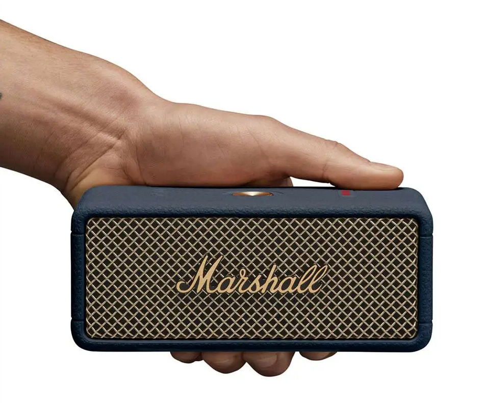 Ręka trzyma niebieski głośnik Marshall ze złotą siatką z przodu. Logo Marshall jest złote. Na górze widoczne jest czerwone światło.
