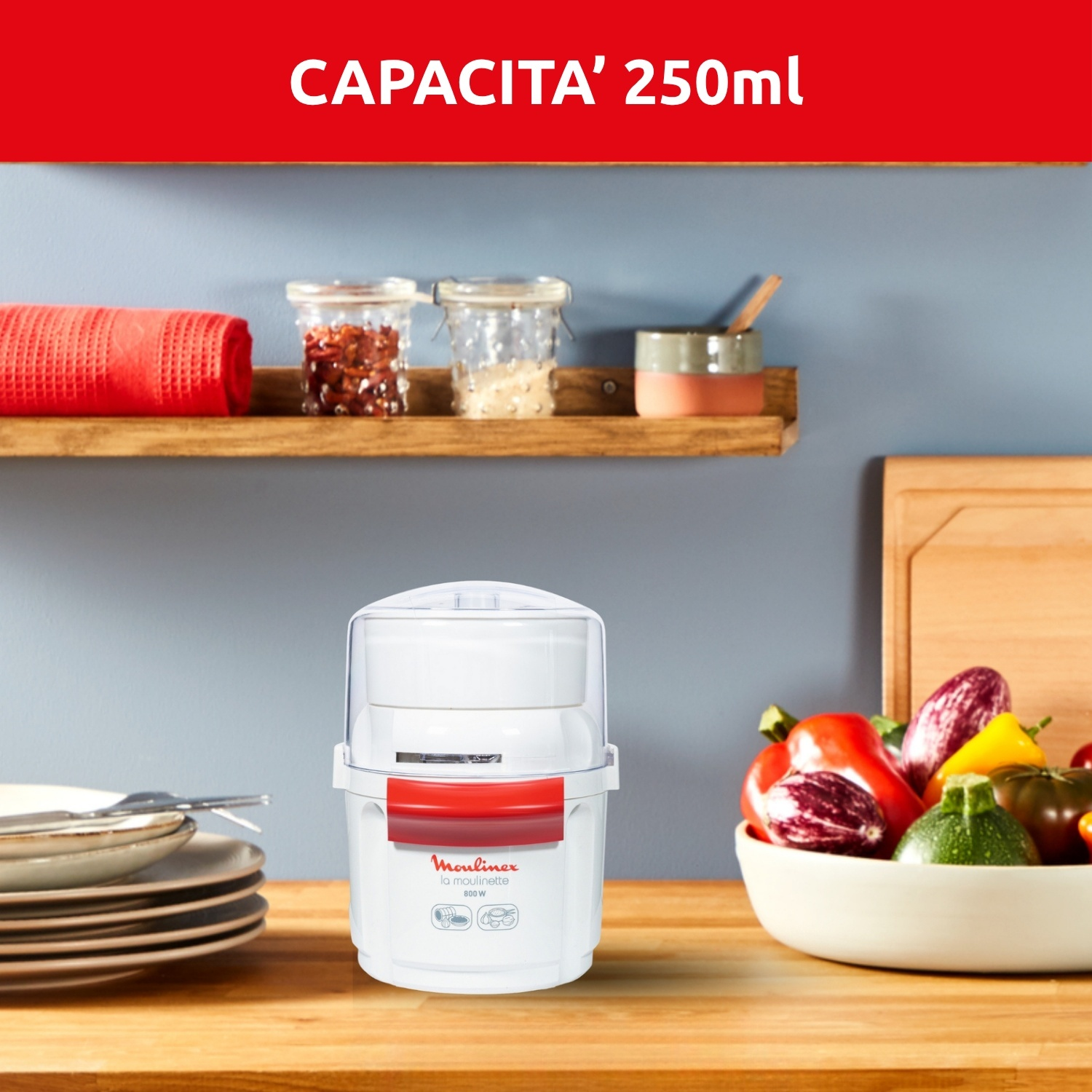 Un robot da cucina Moulinex bianco su un bancone di legno. Verdure in una ciotola e barattoli su uno scaffale sullo sfondo.
