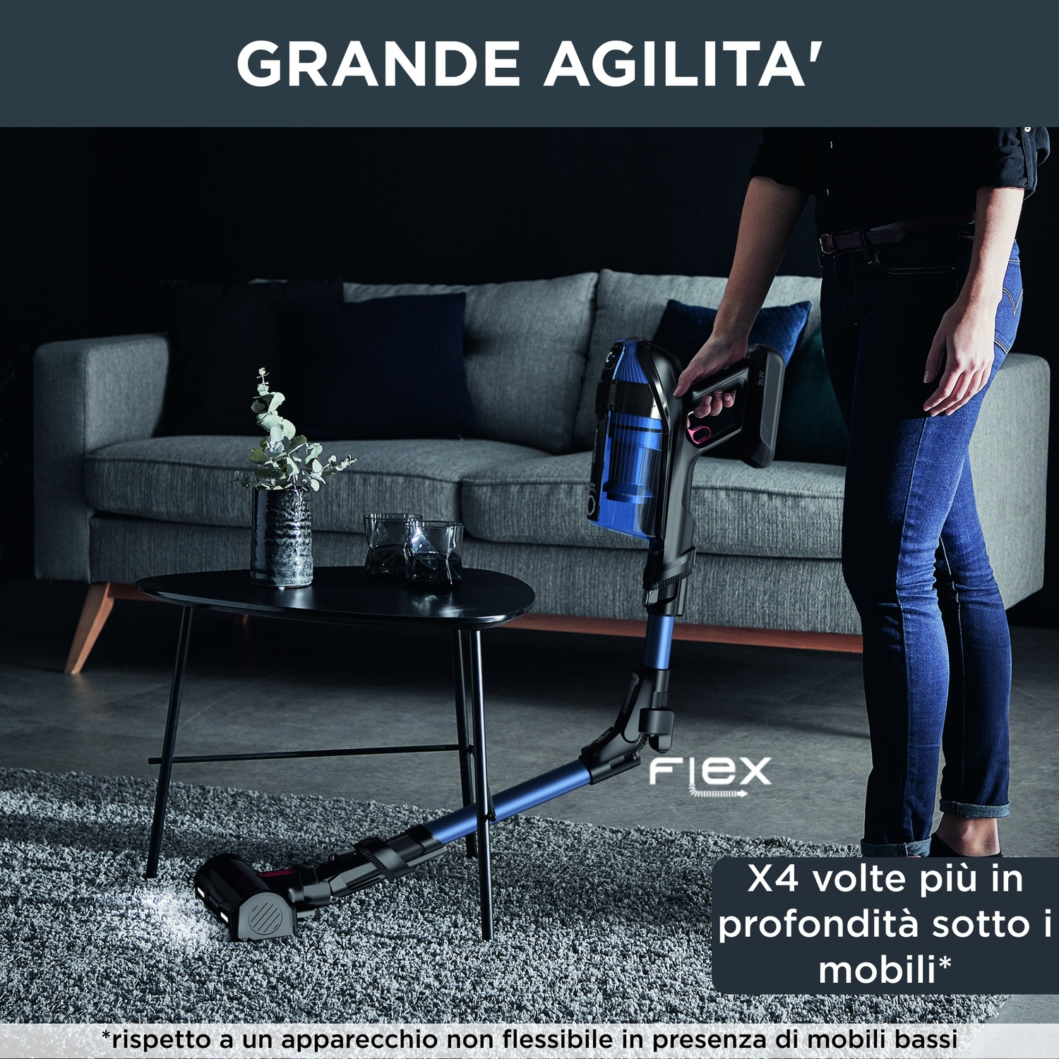 Una donna tiene in mano un aspirapolvere blu e nero. L'aspirapolvere sta pulendo un tappeto grigio. Sullo sfondo un divano e un tavolo.