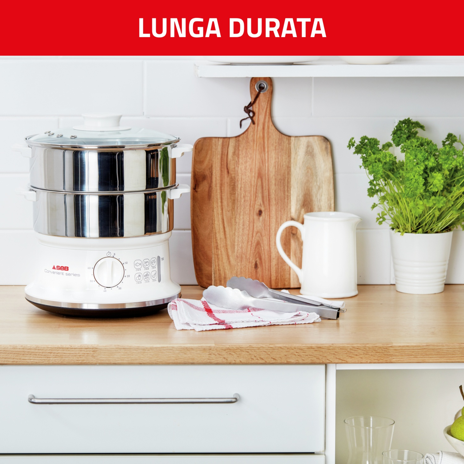 Scena di cucina con vaporiera, tagliere in legno, brocca bianca e pianta. Pinze e un panno sul piano di lavoro.