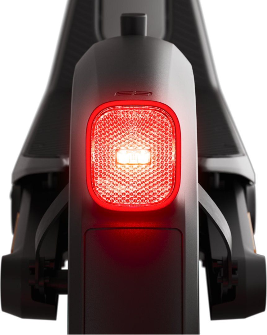 Gros plan de l'arrière d'une trottinette électrique. Un feu arrière rouge vif est central, avec un corps noir et des roues visibles. Fond blanc.