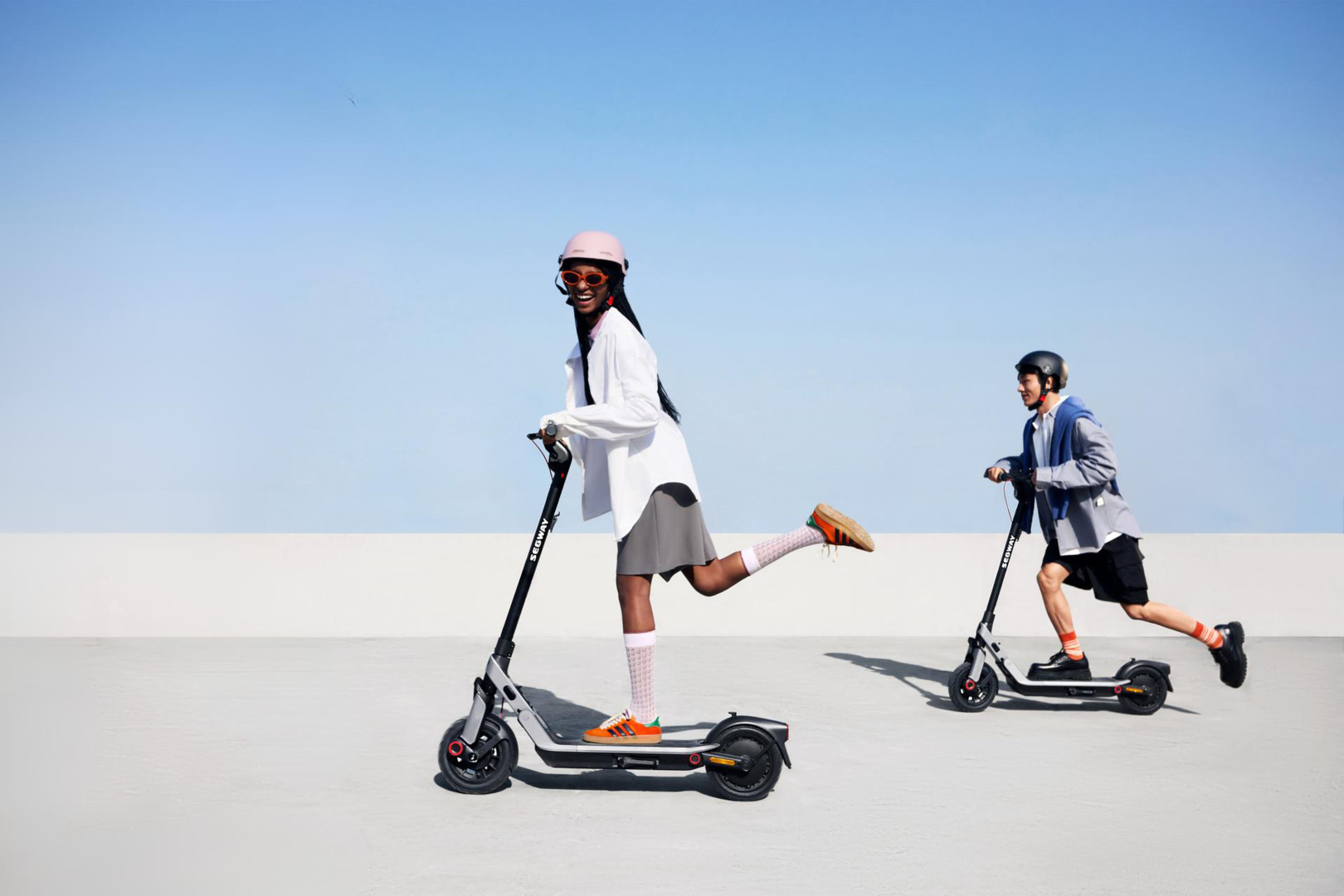 Deux personnes sur des scooters, une en chemise blanche et casque rose, l'autre en veste, contre ciel bleu et surface grise.