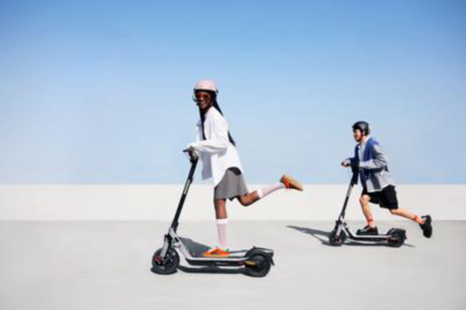 Deux personnes sur des scooters électriques. Une personne porte un casque rose et une chemise blanche. L'autre porte un casque noir et une veste.