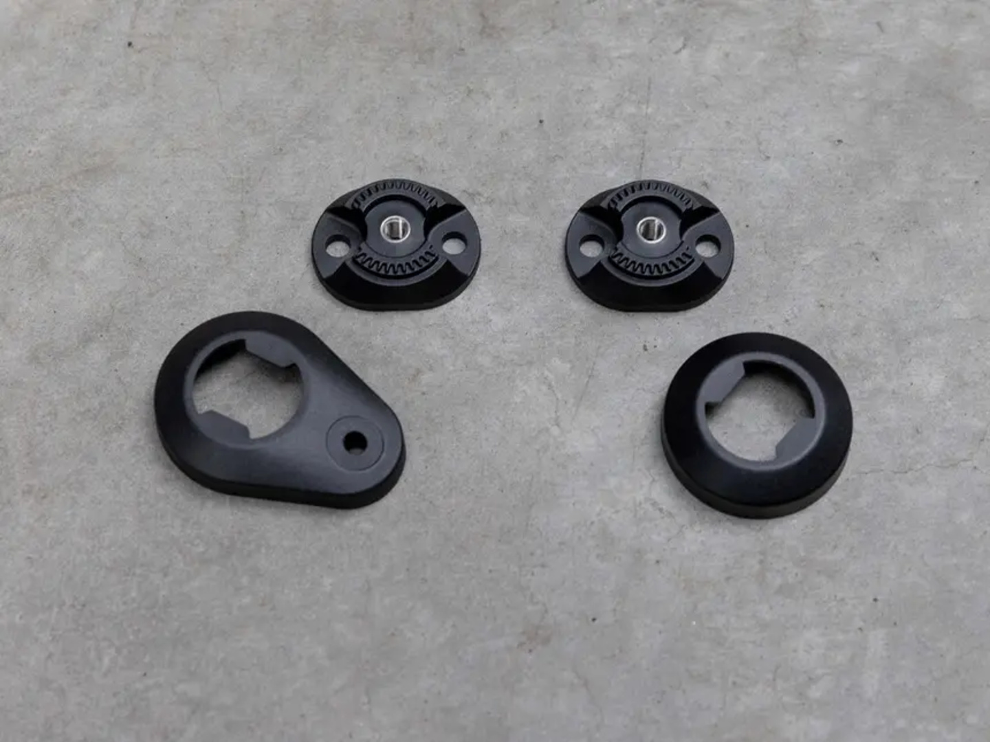 Quattro parti in plastica nera su una superficie grigia. Due sono circolari con fori, una è ovale con un foro e una è un anello.