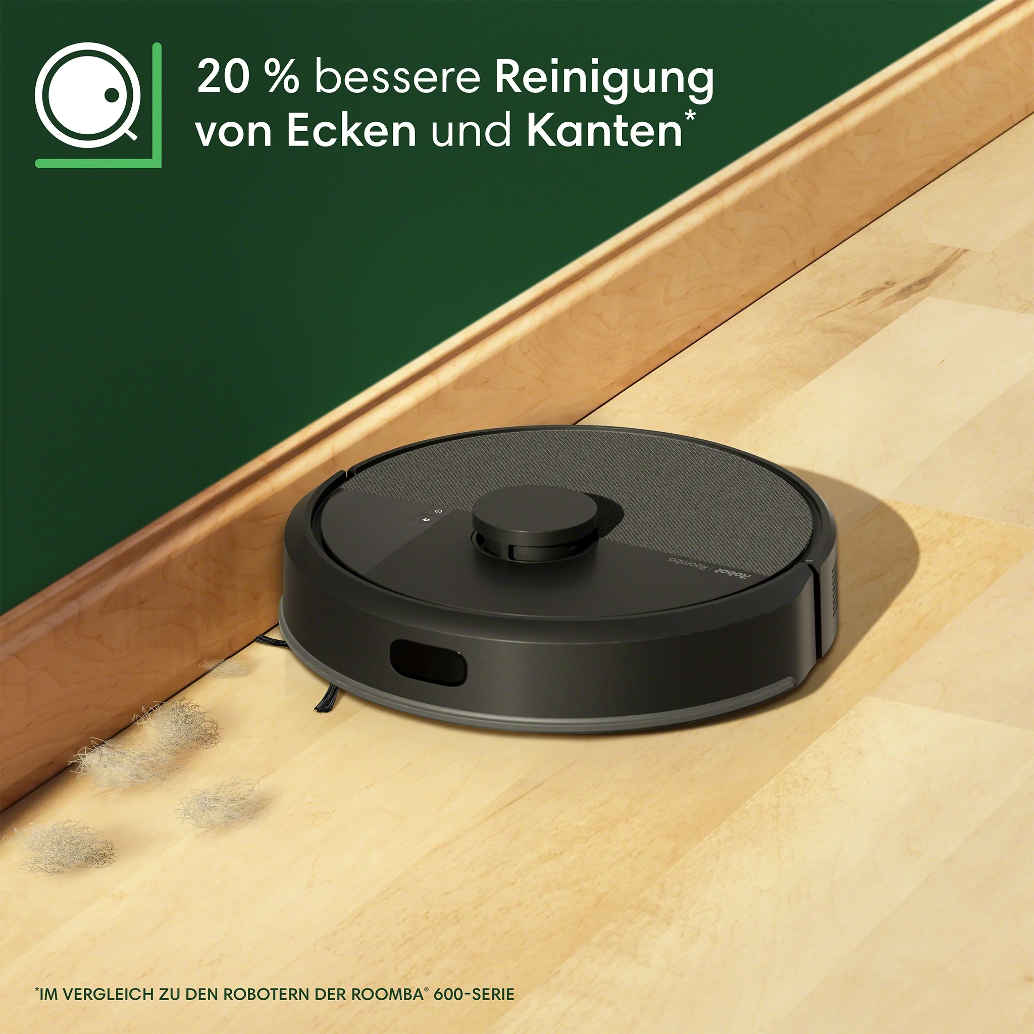 Ein schwarzer Roomba-Staubsauger auf einem Holzboden neben einer Wand. Haare und Text sind sichtbar.