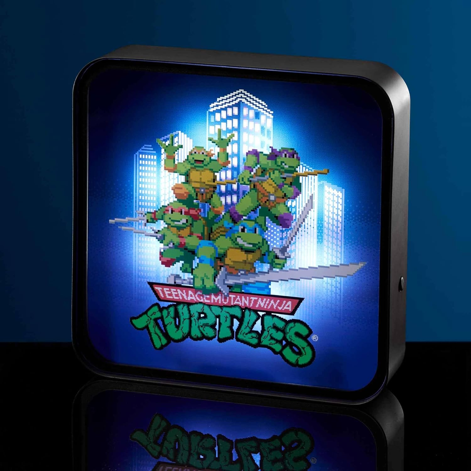 Egy Teenage Mutant Ninja Turtles-es lámpás. A teknősök pixelesek. A háttérben épületek láthatók. A táblán a 'Teenage Mutant Ninja Turtles' felirat szerepel.