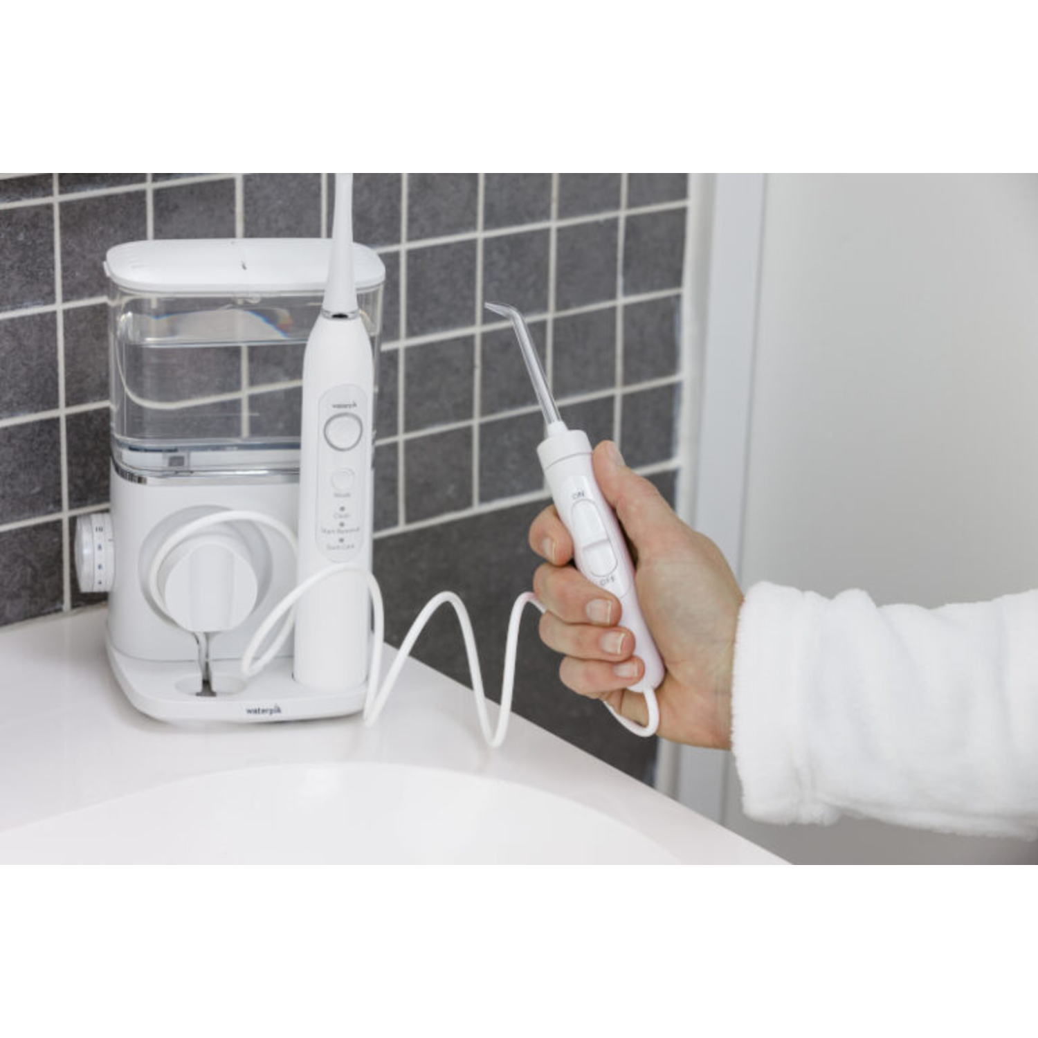 Een persoon houdt een witte waterflosser vast. Een andere flosser staat op het aanrecht. De achtergrond is een witte badkamer.