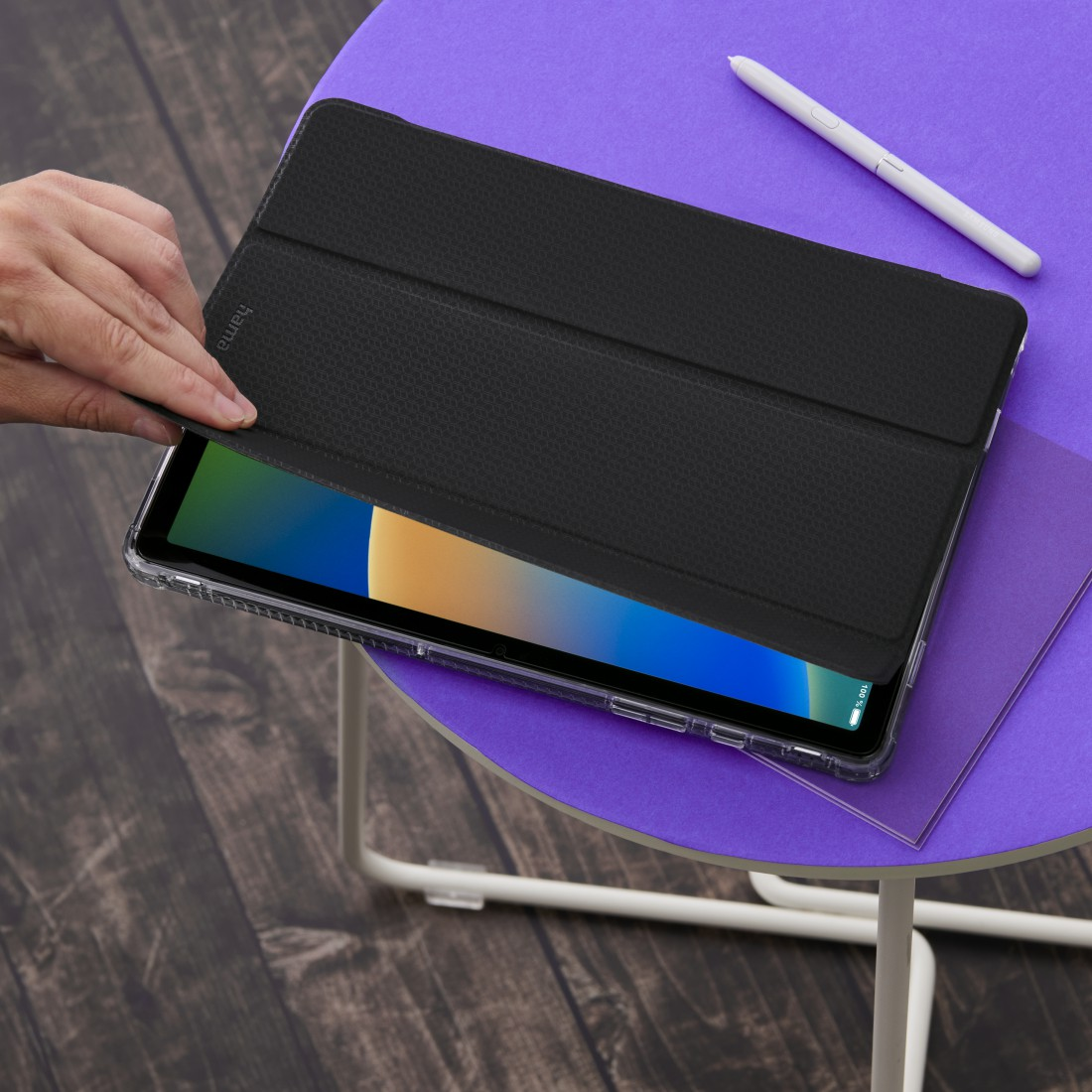 Eine Hand öffnet eine schwarze Tablethülle auf einem lila Tisch. Ein weißer Stift und ein lila Papier liegen ebenfalls auf dem Tisch.