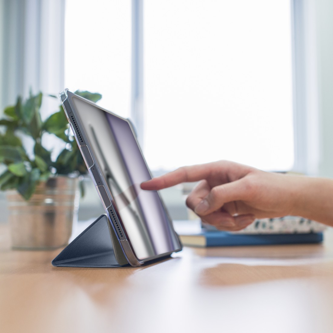 Eine Hand berührt einen Tablet-Bildschirm. Das Tablet befindet sich in einem Gehäuse und ist aufgestellt. Im Hintergrund befinden sich eine Pflanze und Bücher.