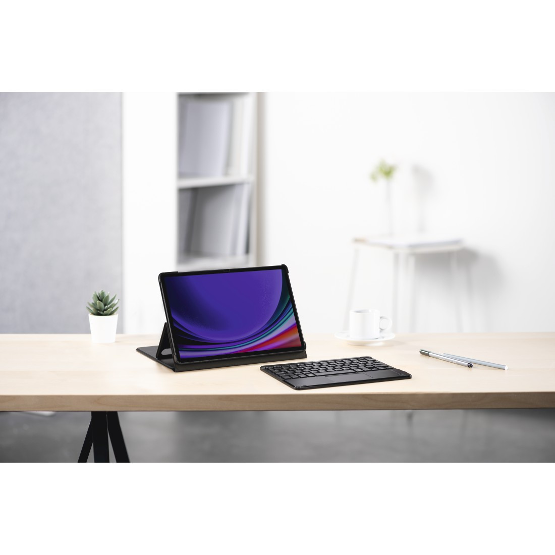 Tablet mit Tastatur auf einem Holztisch. Links und rechts eine kleine Pflanze und eine Tasse. Hintergrund ist eine weiße Wand.