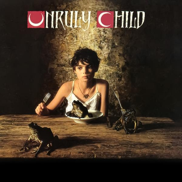 Eine junge Frau sitzt an einem Holztisch mit Fröschen. Ein Frosch ist in einer Schüssel. Der Text oben lautet 'Unruly Child'.