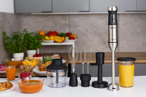 Scena kuchenna z blenderem ręcznym, akcesoriami i produktami spożywczymi, takimi jak zupa i sok na białej powierzchni.